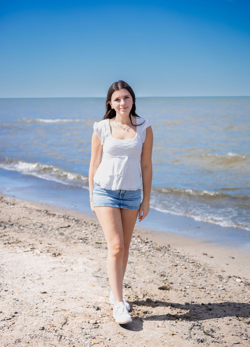 Young woman in white top and denim shorts walking on a sandy beach with calm ocean waves under a clear blue sky.