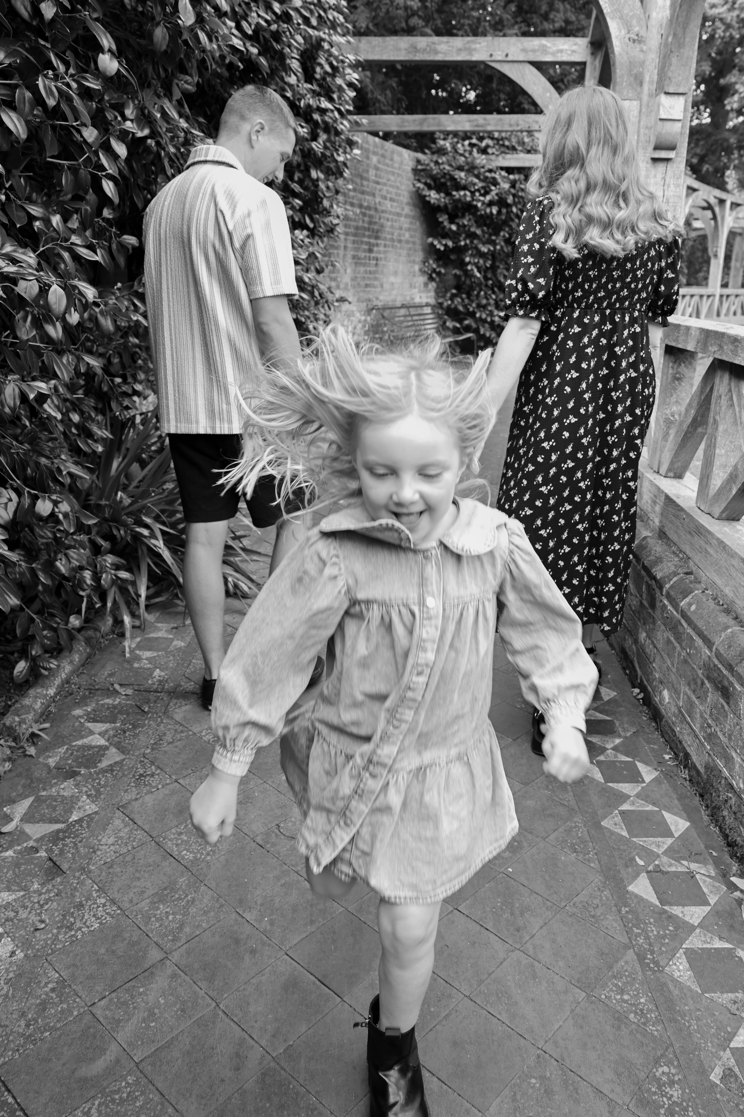 Black and white photo of a joyful girl with windswept hair running ahead of two adults on a garden path.