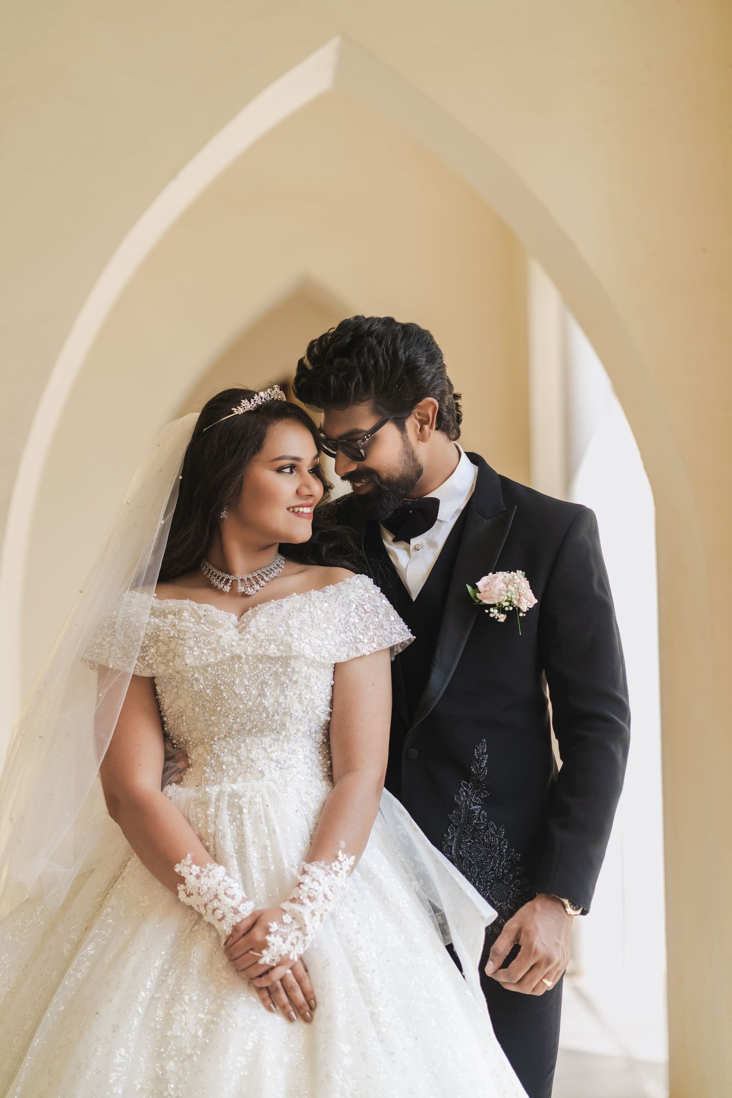 A bride in a white ball gown and groom in a black tuxedo share a romantic moment under an arch.