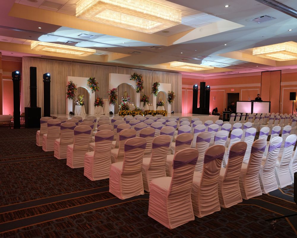 Elegant wedding ceremony venue with white chair covers, purple sashes, floral backdrop, and warm pink uplighting in a ballroom.