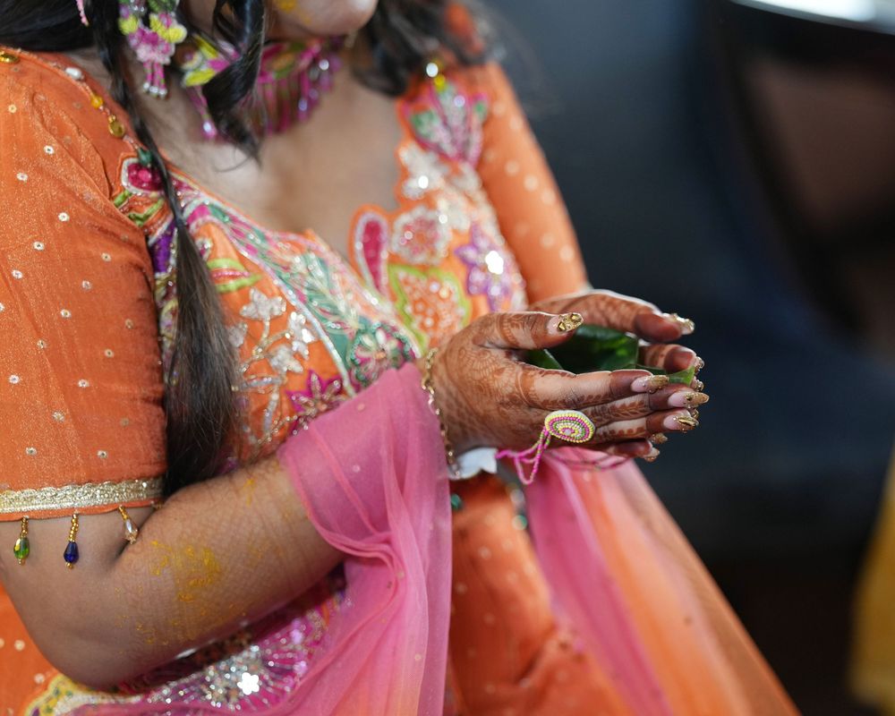Woman in vibrant orange and pink Indian bridal attire with mehndi hands holding green leaves at a ceremony.
