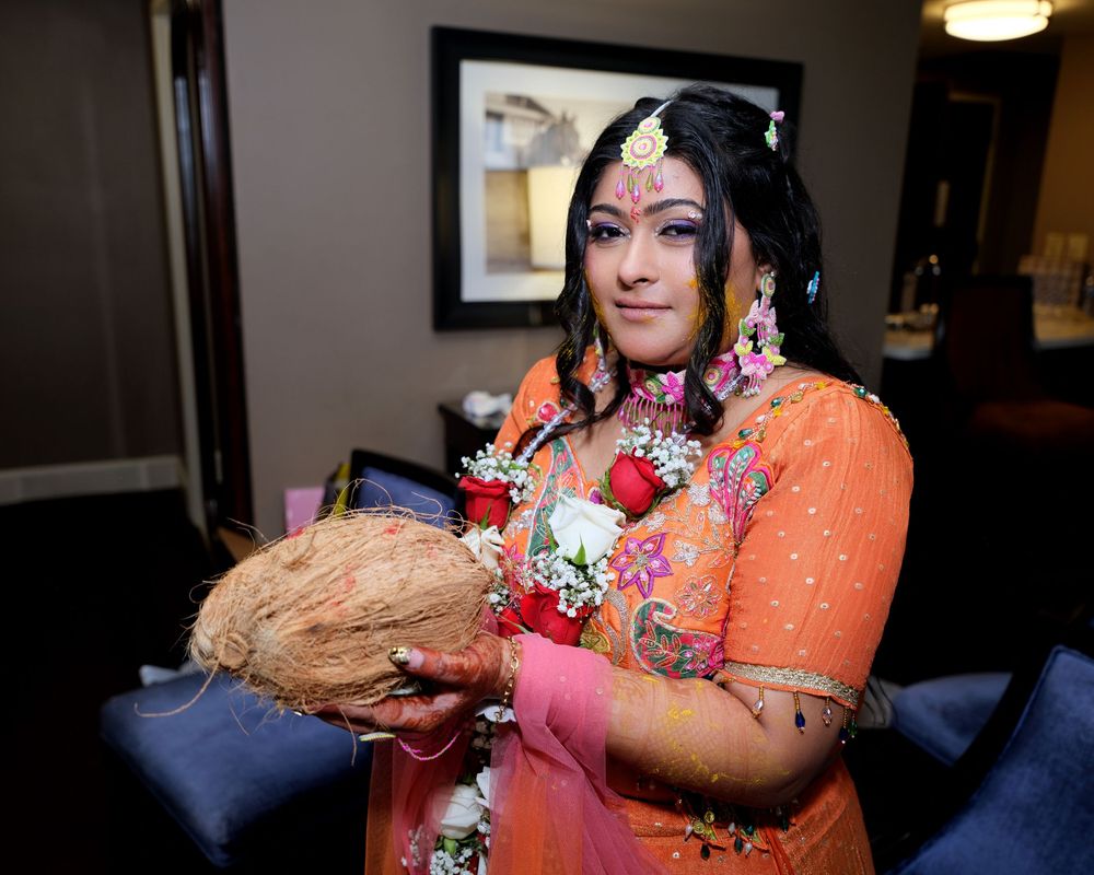 Indian bride in orange salwar kameez holding a coconut, adorned with traditional jewelry and floral garlands.