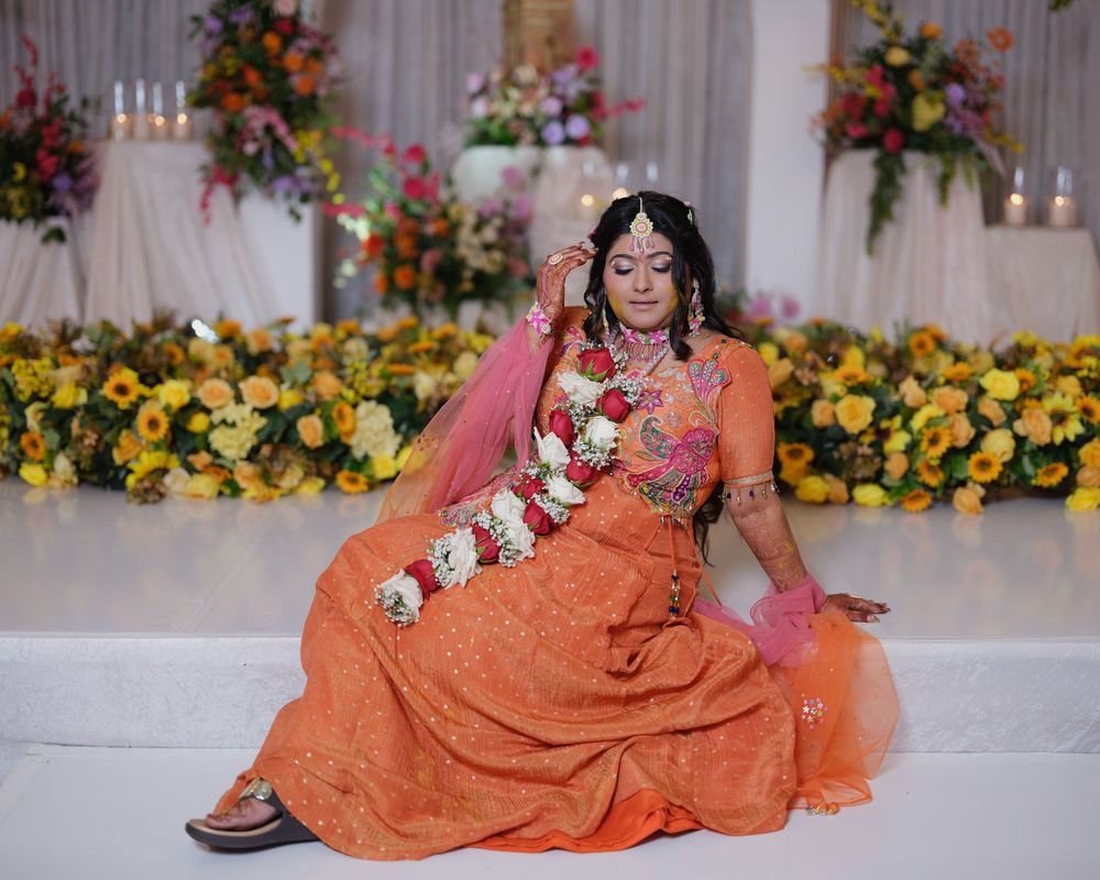 Bride in orange lehenga with floral garland seated gracefully at decorated Indian wedding venue.