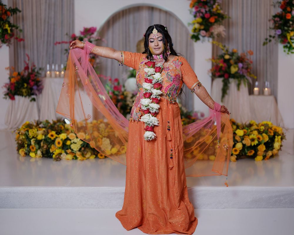 Woman in orange traditional dress with floral garland posing gracefully at decorated venue.