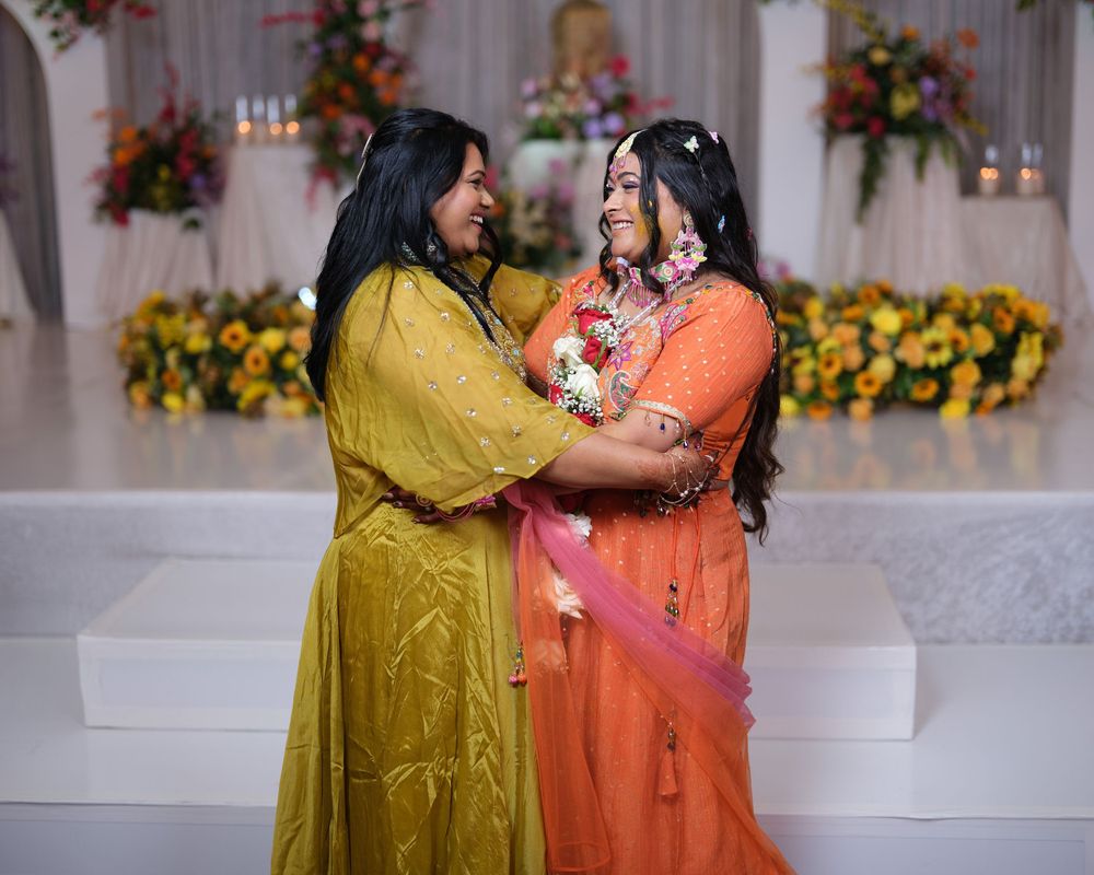 Two women in vibrant yellow and orange Indian attire share a joyful embrace at a decorated wedding ceremony.