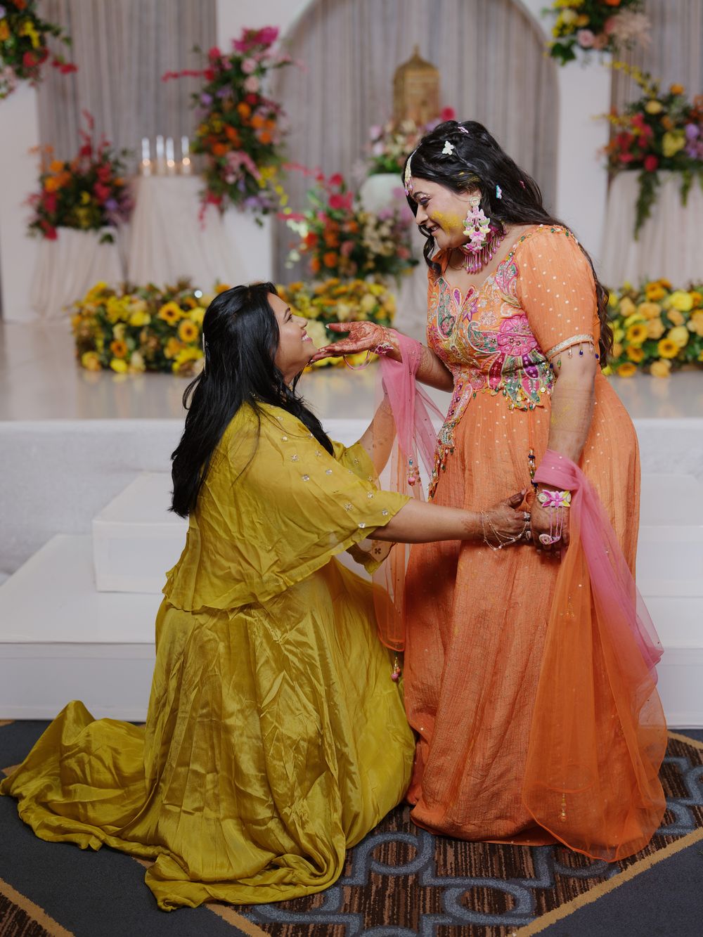 Two women in vibrant Indian attire share a joyful moment at a decorated wedding ceremony venue.