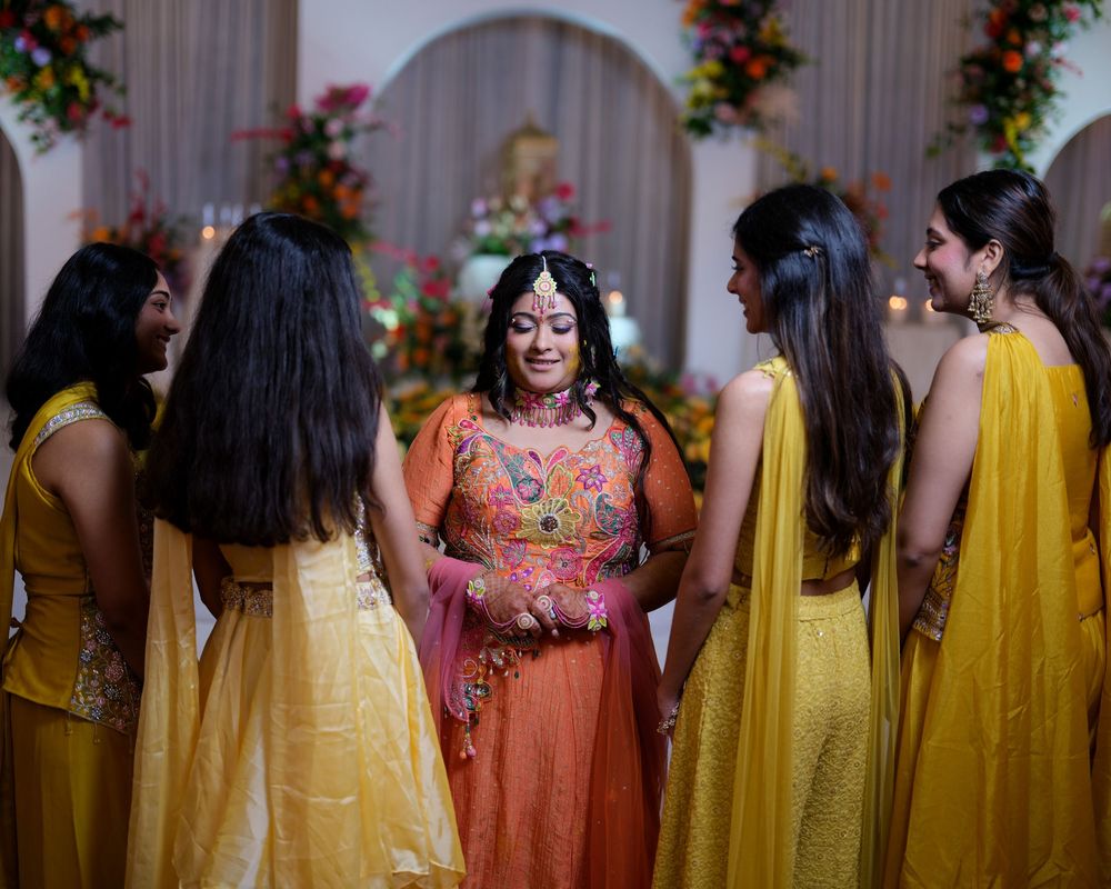 Indian bride in orange lehenga surrounded by bridesmaids in yellow outfits during a traditional wedding ceremony.