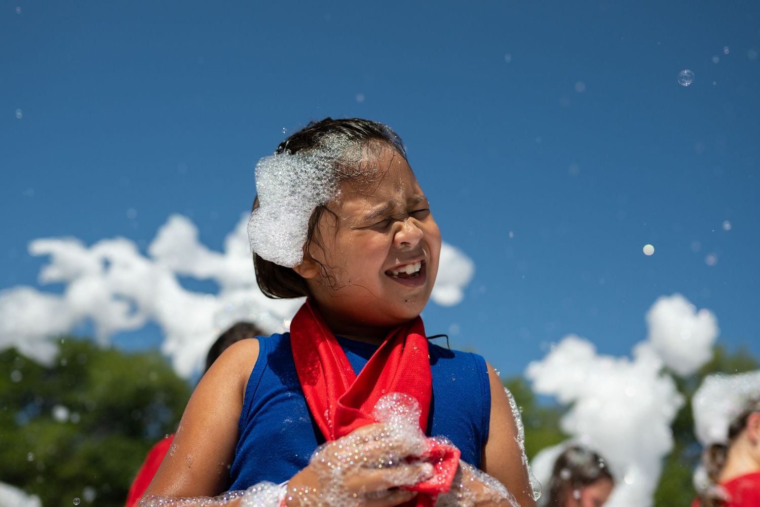 Child covered in foam bubbles playing outdoors on a sunny day with blue sky and clouds in the background.