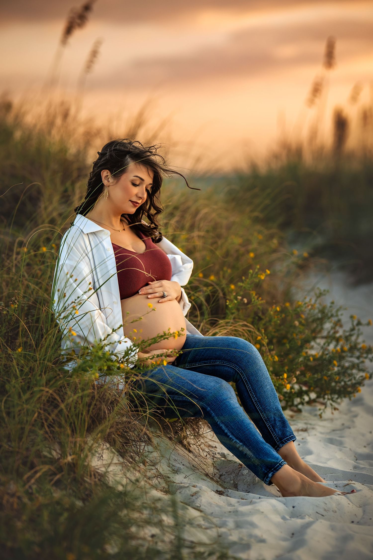 Pregnant woman sitting on beach sand dunes at sunset, cradling her baby bump, wearing jeans and open white shirt.