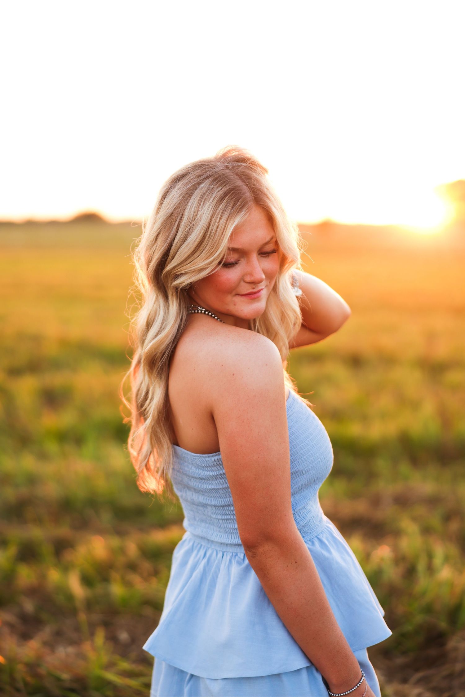 Blonde woman in blue dress smiling at golden sunset in open field.