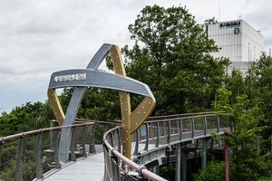 Modern pedestrian bridge with golden arch and metal railings winds through lush green trees near a white building.