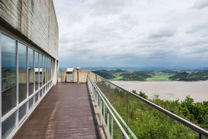 A modern building walkway with wooden deck and glass windows overlooking a scenic river valley under cloudy skies.