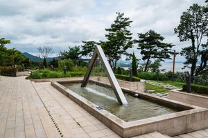 Modern fountain with triangular sculpture in a stone plaza surrounded by lush trees and mountain views under cloudy skies.