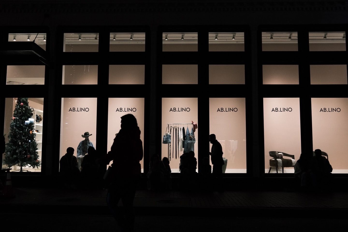 Silhouettes of shoppers walk past illuminated AR.UNO store windows at night during the holiday season.