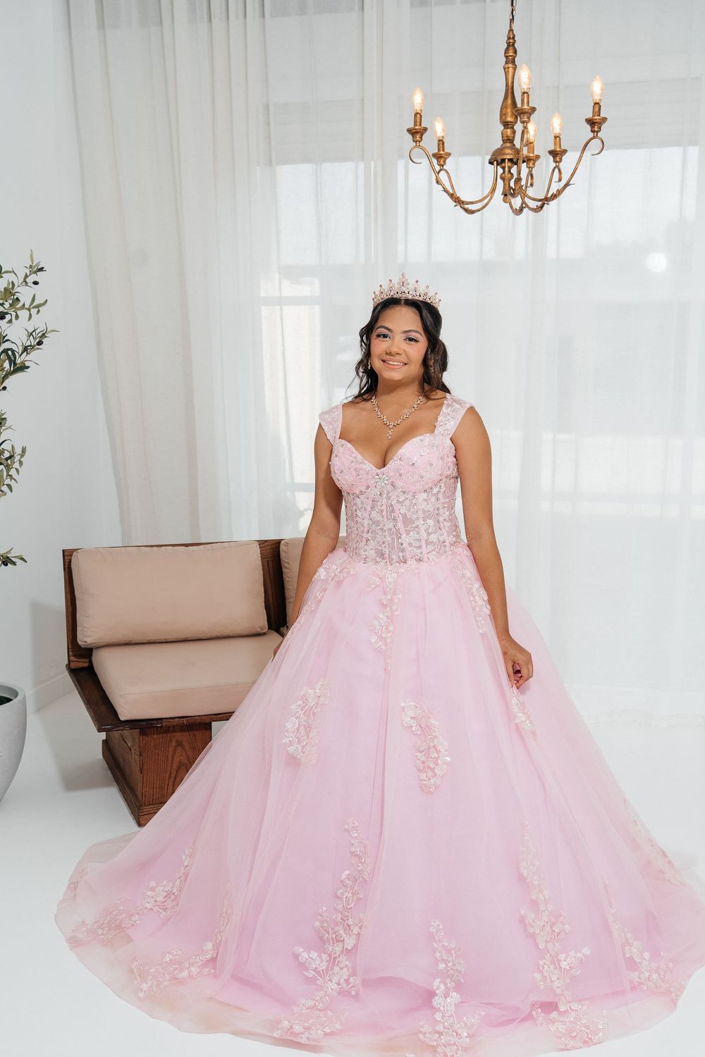 Young woman wearing a pink ball gown with floral lace details and a tiara, posing in a bright white studio.