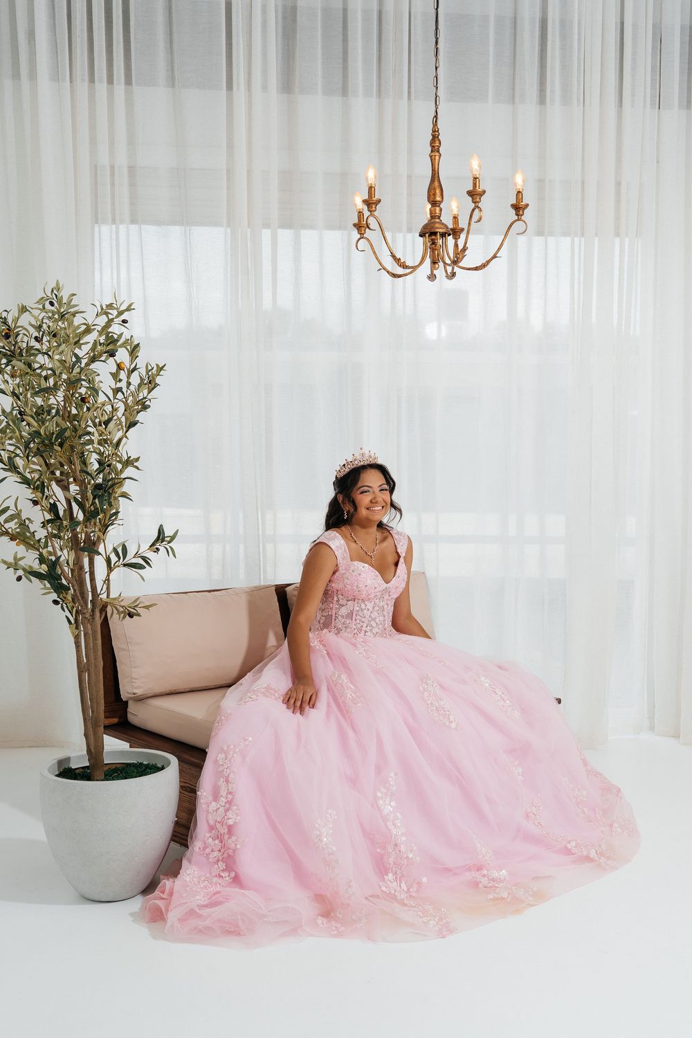 Young woman in pink floral ball gown with crown sits gracefully on a bench beneath a gold chandelier.