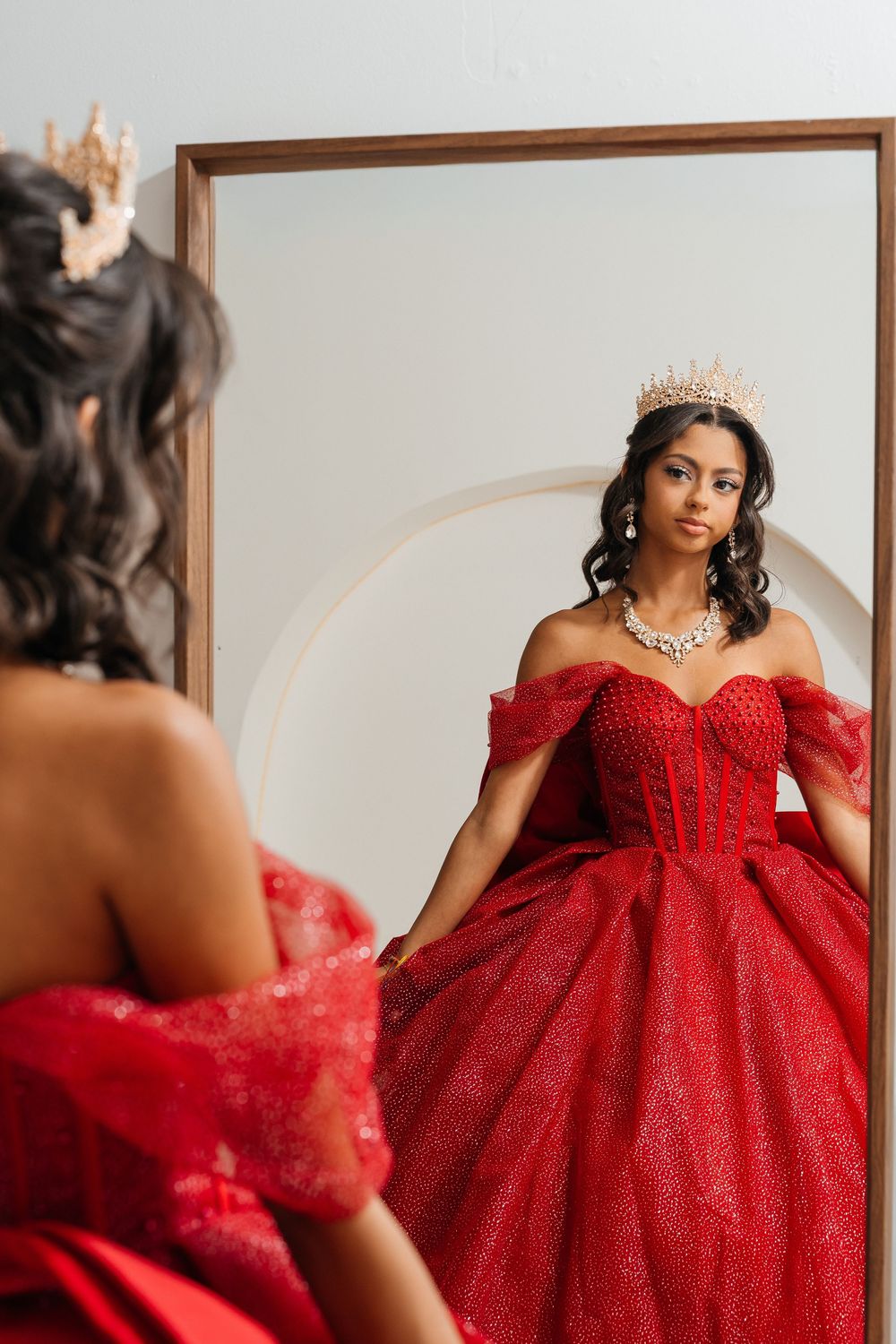 Young woman in red off-shoulder ballgown and gold crown gazes at her reflection in a large mirror.
