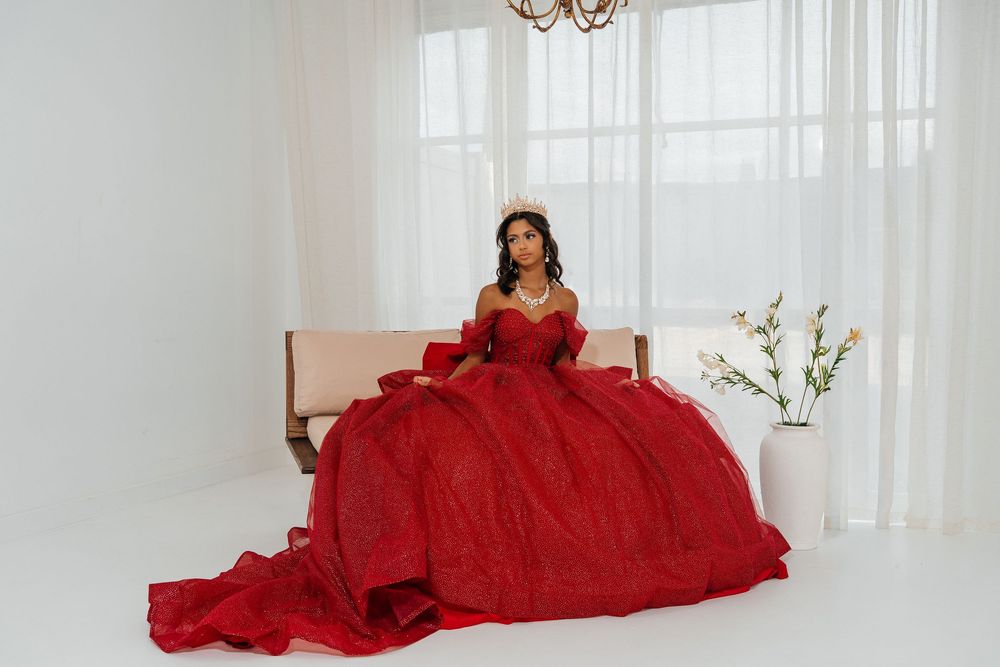 Young woman in a stunning red ball gown with gloves and tiara poses elegantly in a bright white room.