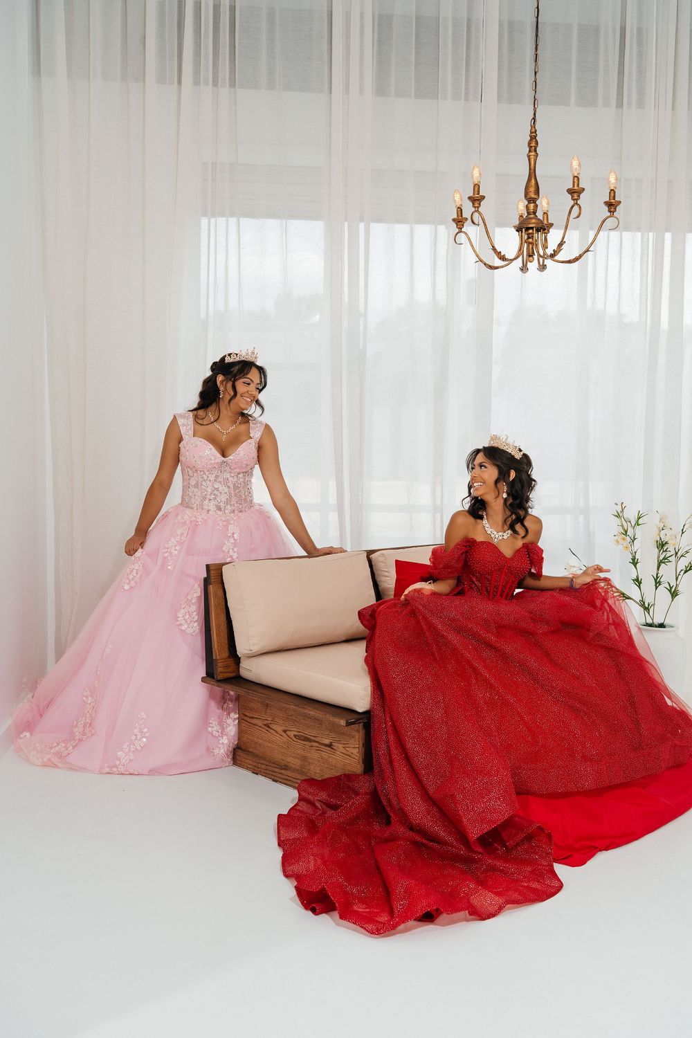 Two women in elegant ball gowns, one pink and one red, pose in a bright white room with a chandelier.