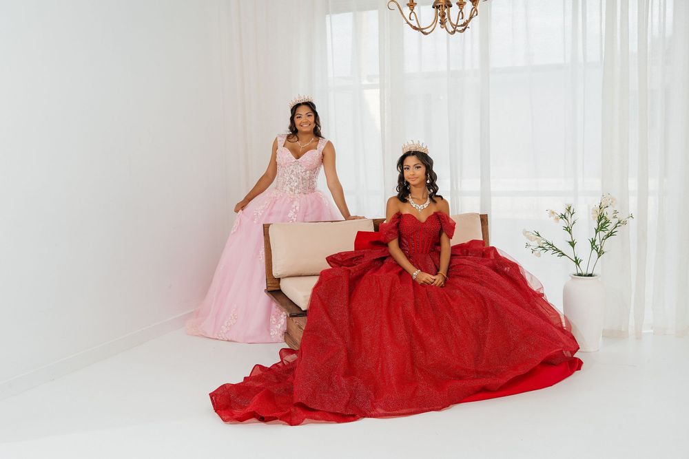 Two women in elegant ball gowns, one pink and one red, pose in a bright white studio setting.