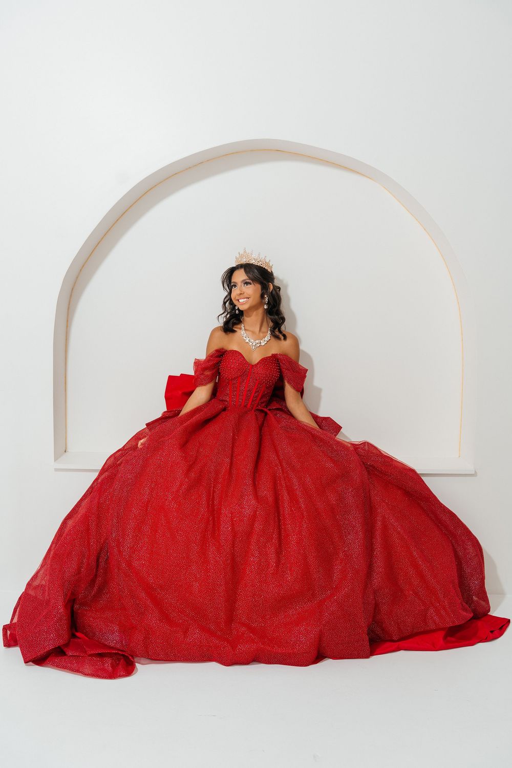 Young woman in a stunning red off-shoulder ball gown with a tiara poses gracefully against a white arched backdrop.