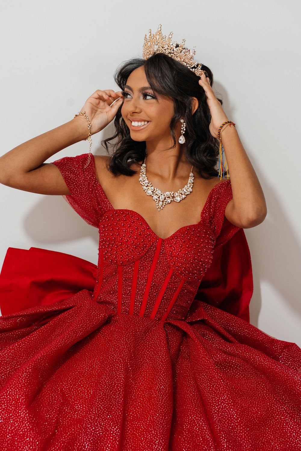 Young woman in red sparkly ball gown with gold crown and diamond necklace smiling elegantly.