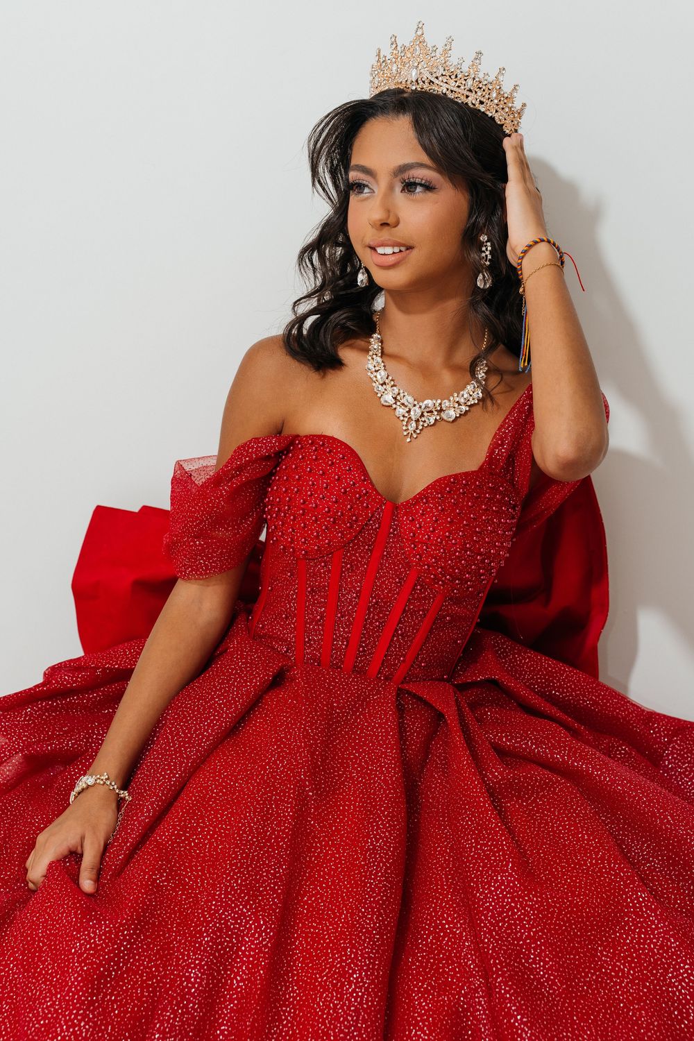 Young woman in a red sparkly off-shoulder ball gown with a gold crown and diamond jewelry poses elegantly.