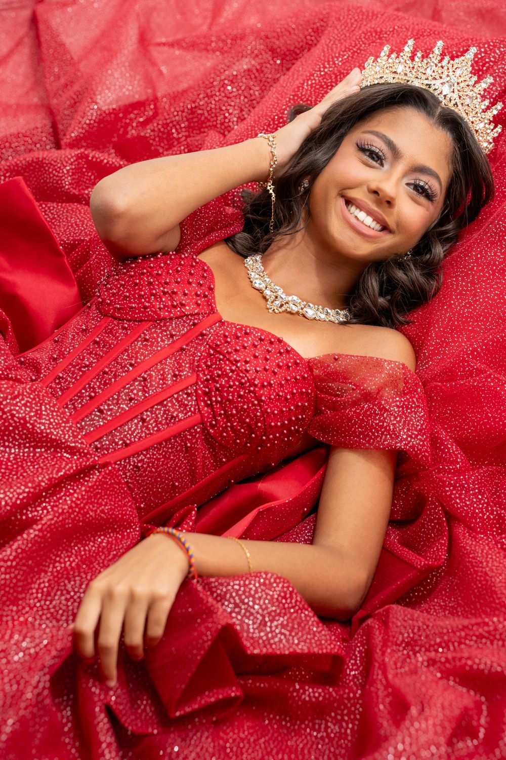 Young woman in red beaded off-shoulder quinceañera gown and crown smiles while posing on red sparkly fabric.