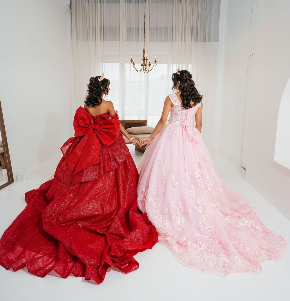 Two women in elegant ballgowns, one red and one pink, holding hands while facing a window in a bright room.
