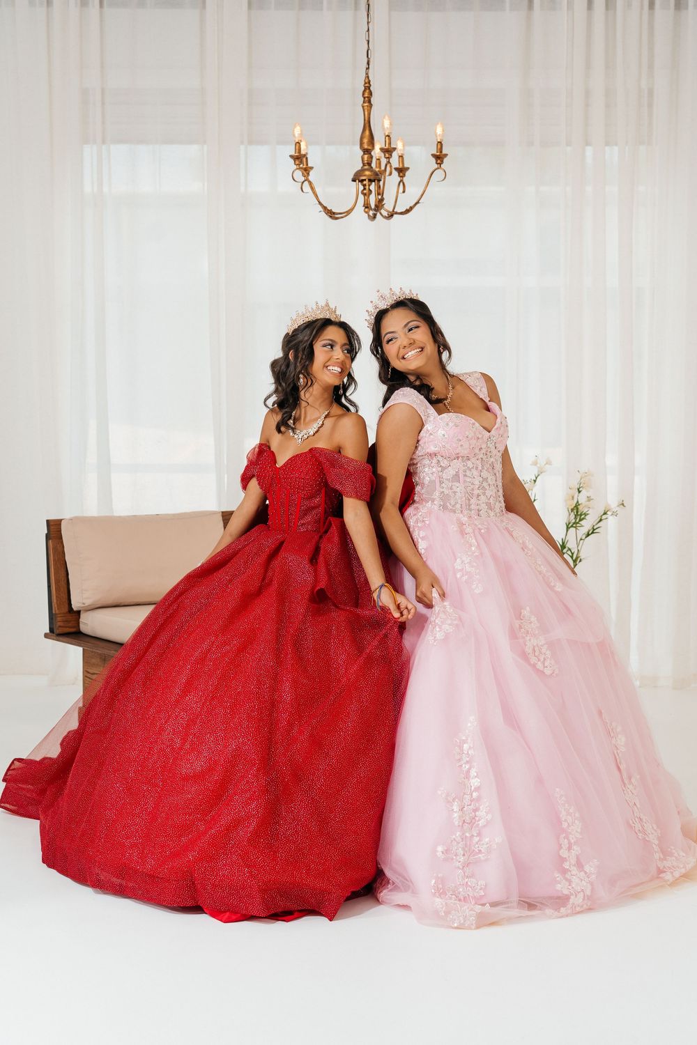 Two young women in red and pink ball gowns with crowns pose together in elegant white room.