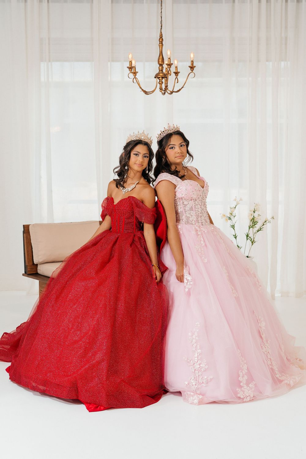 Two women in red and pink ball gowns with crowns pose elegantly in a bright white studio setting.