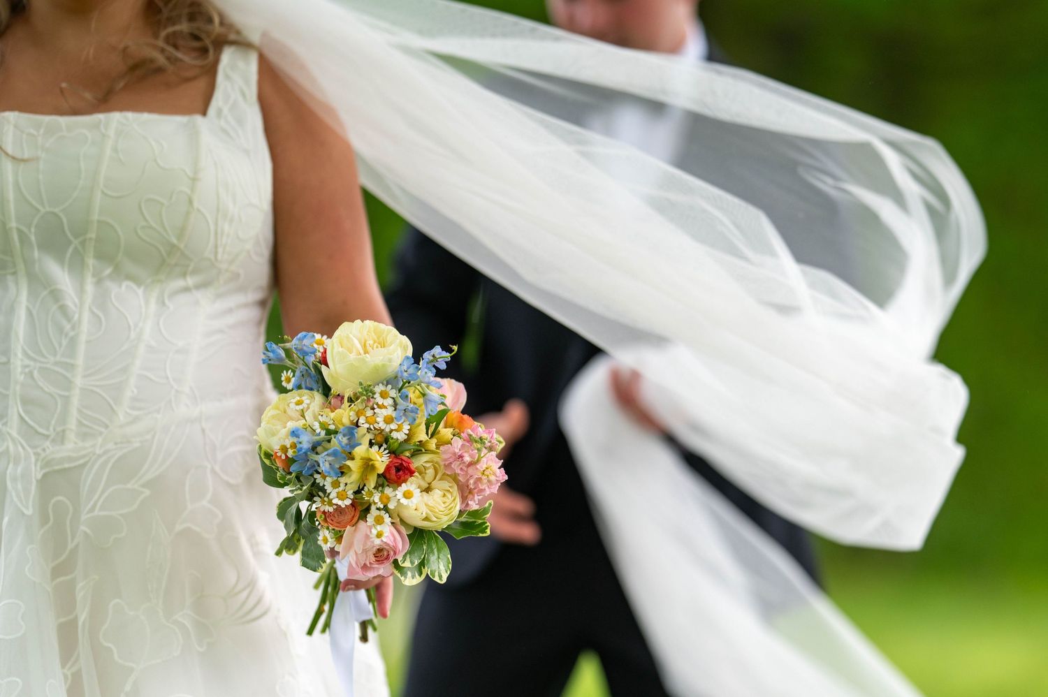 Bride holding colorful spring bouquet with veil blowing in wind beside groom in black suit outdoors.