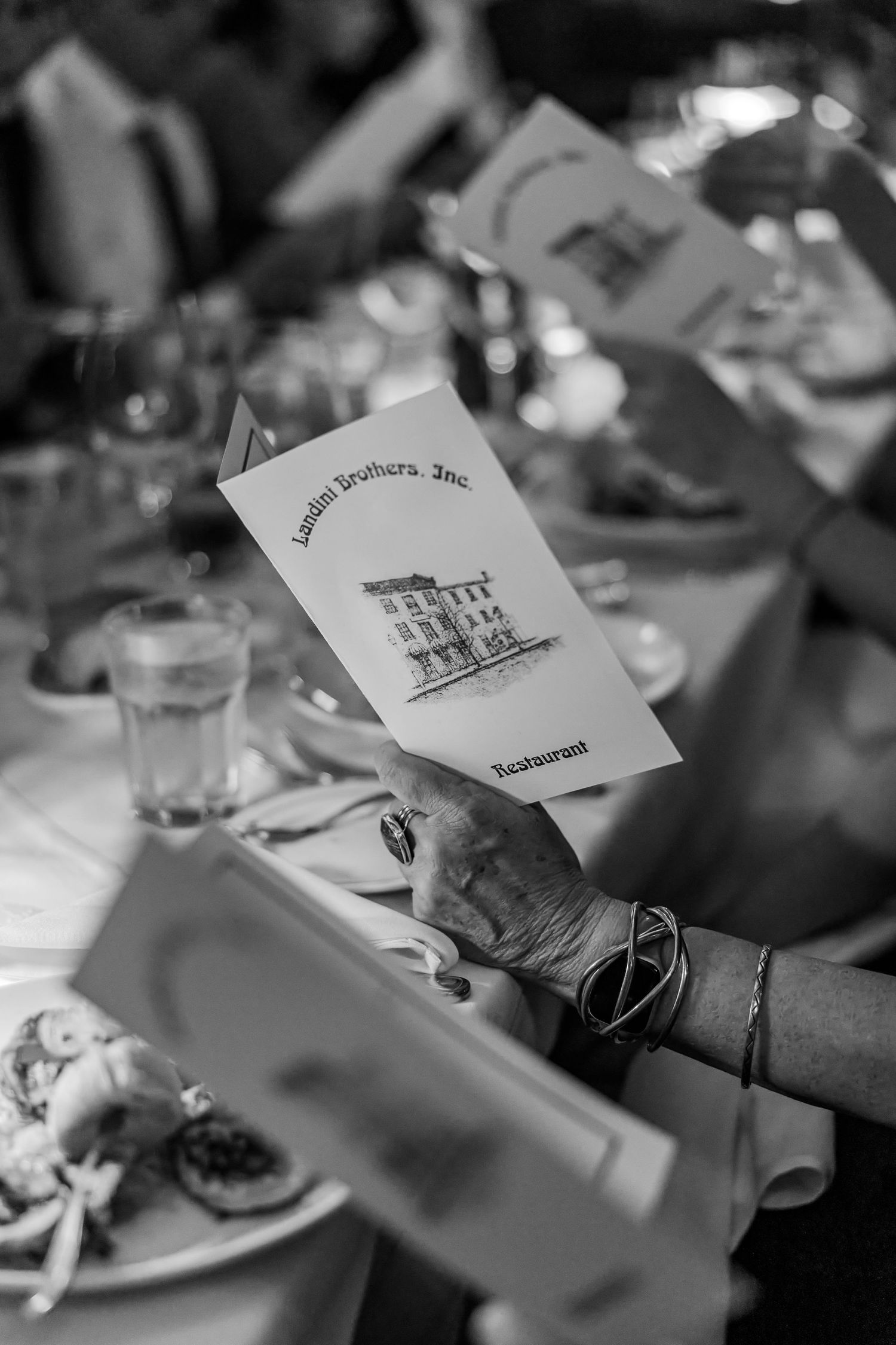 Black and white photo of a hand holding a Landini Brothers Inc. Restaurant menu at a formal dining table.