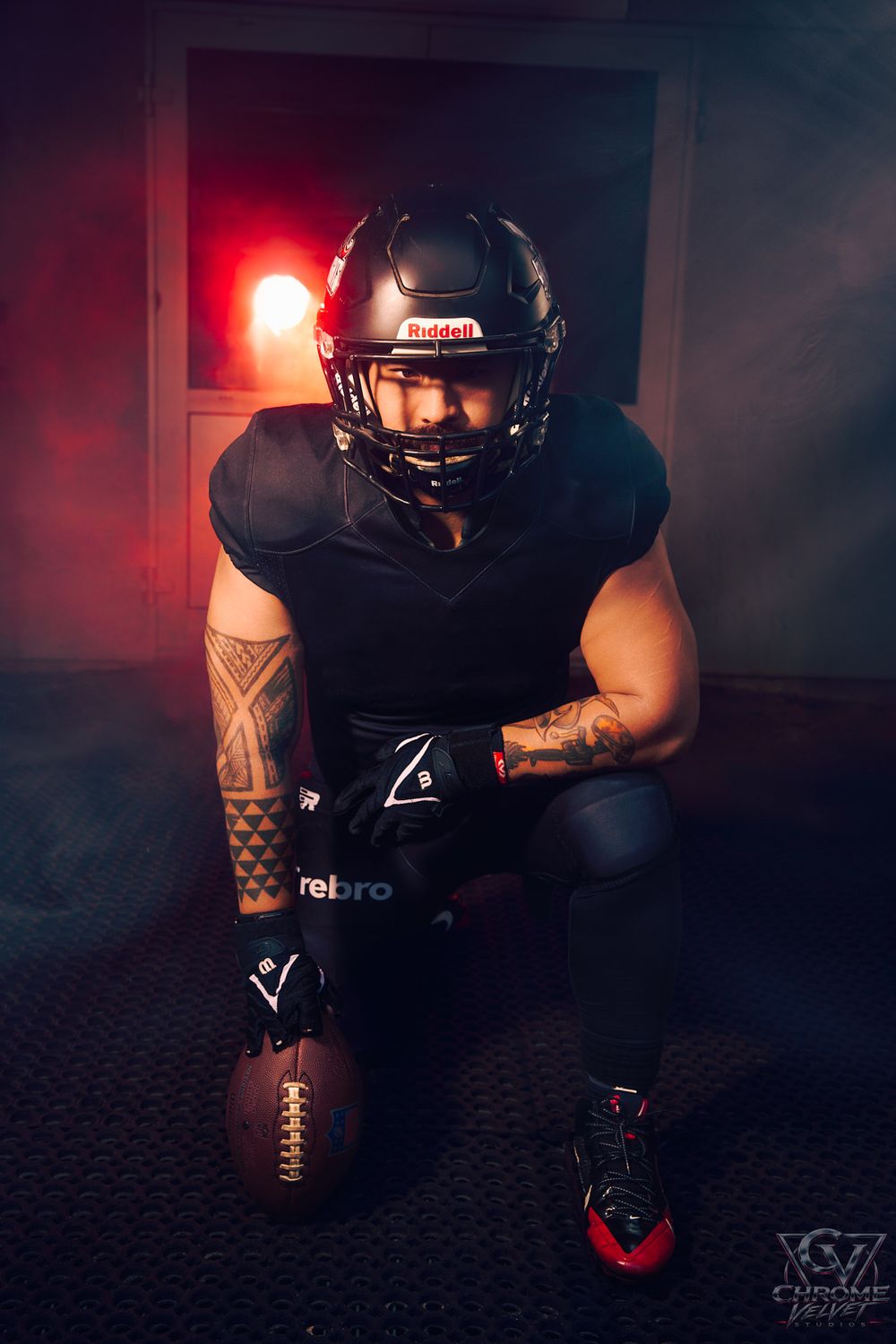 A tattooed football player kneels in a dark, atmospheric studio while gripping a football, wearing black protective gear, a black helmet, gl