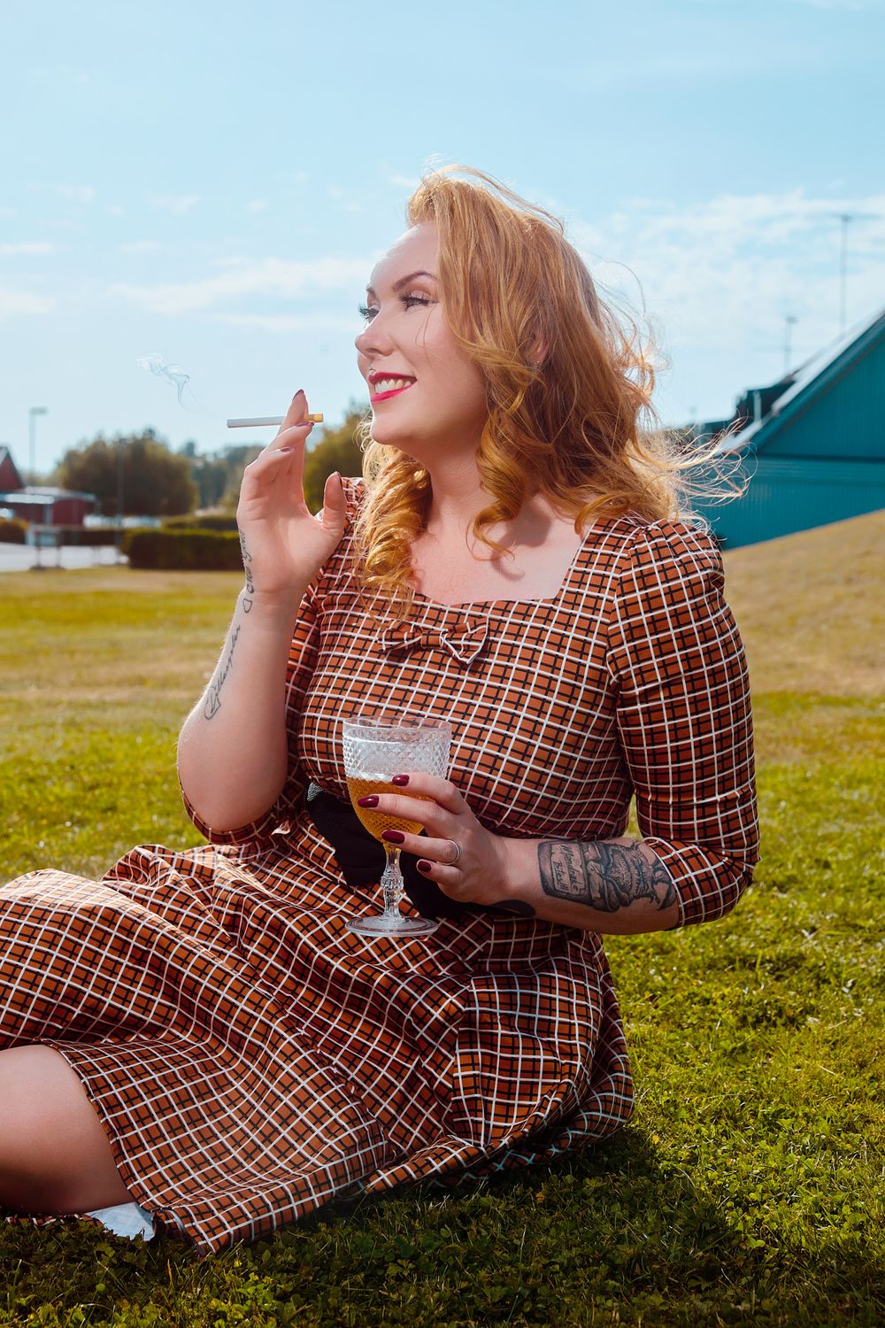 Retro rockabilly portrait of a tattooed red-haired woman sitting on grass in a vintage plaid dress, holding a cocktail glass and cigarette w