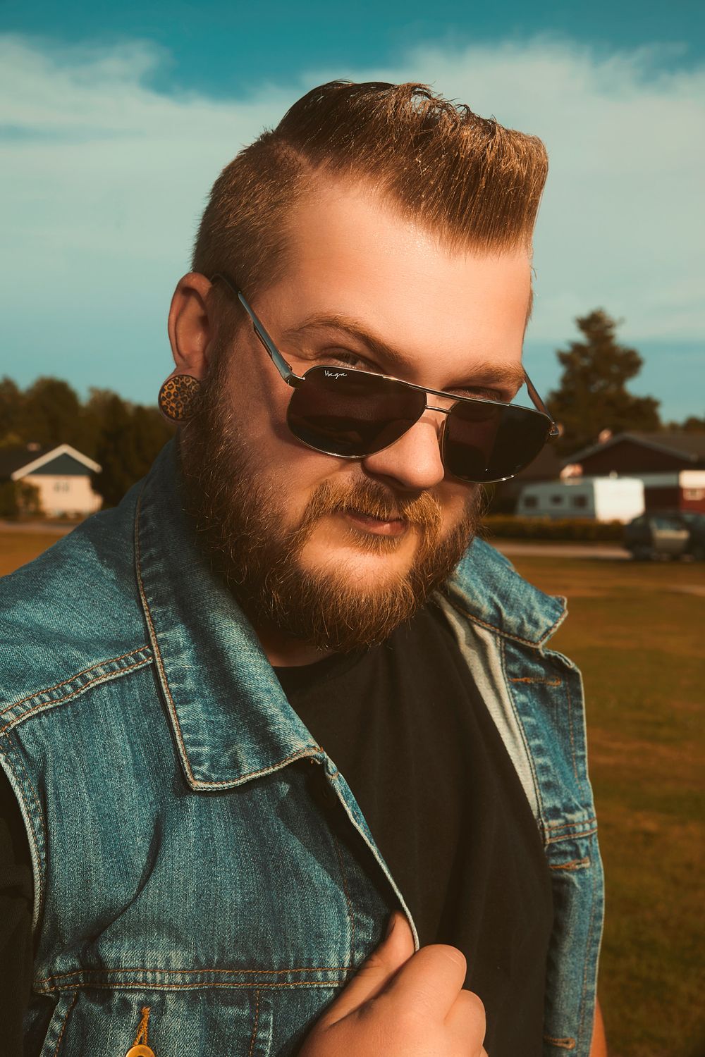 Close-up portrait of a bearded man with styled pompadour hair, wearing dark vintage sunglasses, a denim jacket, black shirt, and large patte
