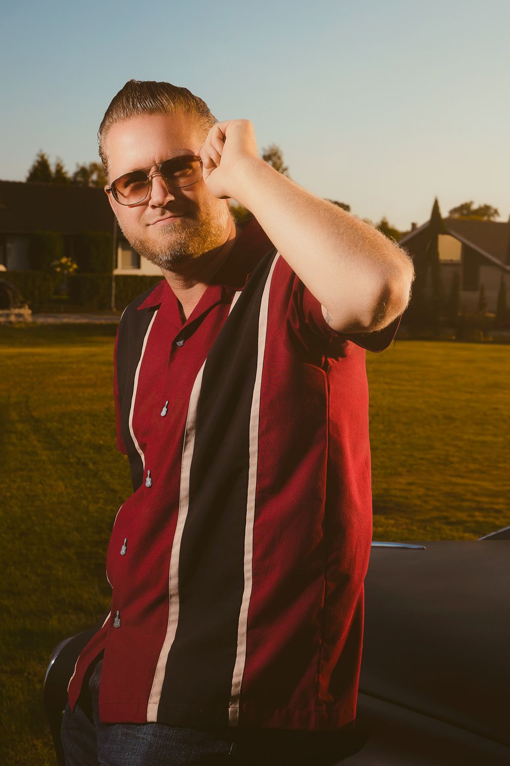 Portrait of a man with slicked-back pompadour hair and sunglasses wearing a red and black vintage bowling shirt with white stripes, posing b
