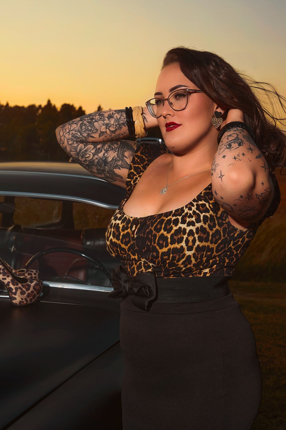 Portrait of a tattooed woman wearing black cat-eye glasses, a leopard print top, and black pencil skirt, posing beside a classic vintage car