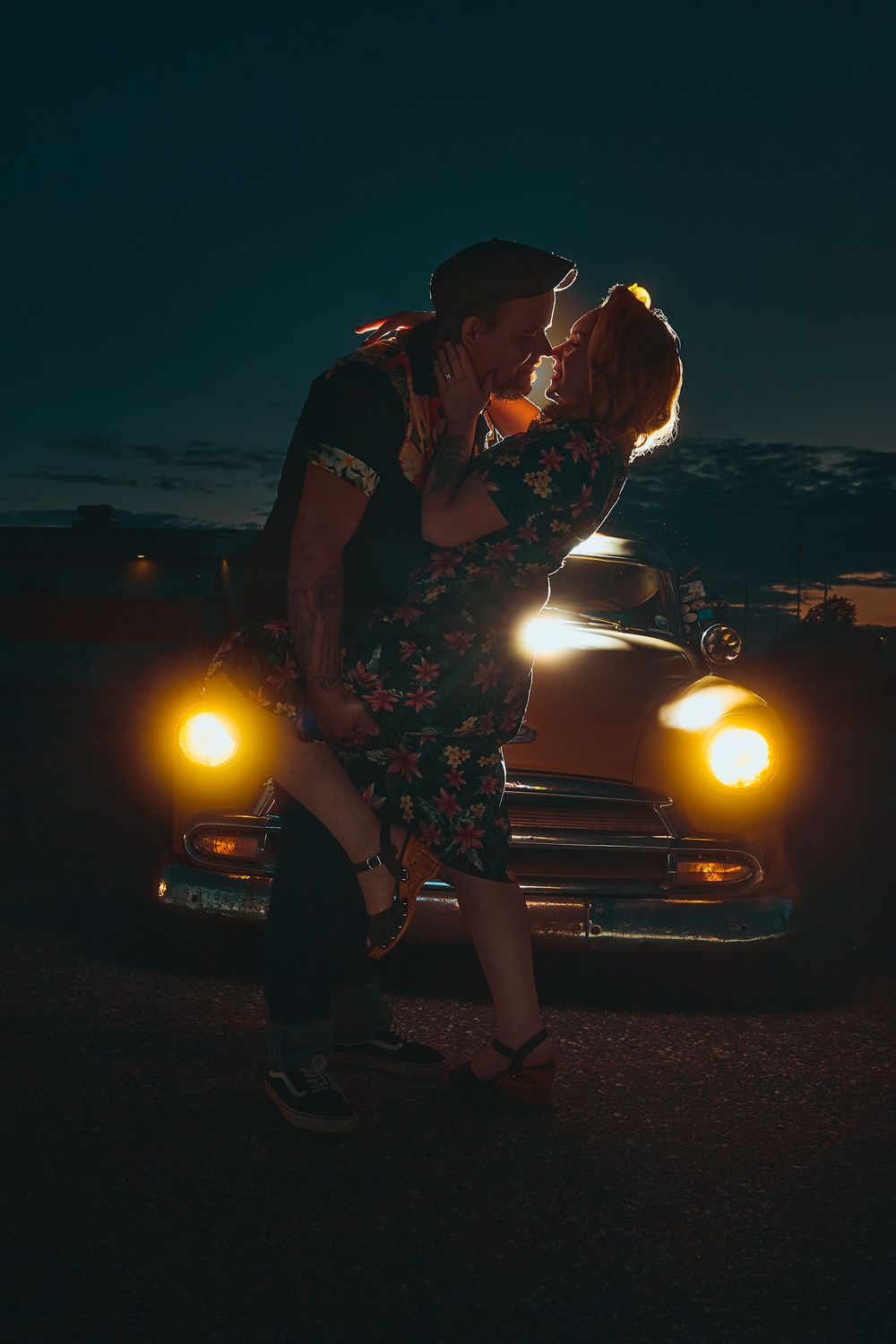 Retro rockabilly couple embracing and kissing in front of a classic vintage car at dusk, illuminated by the car’s glowing headlights, with t