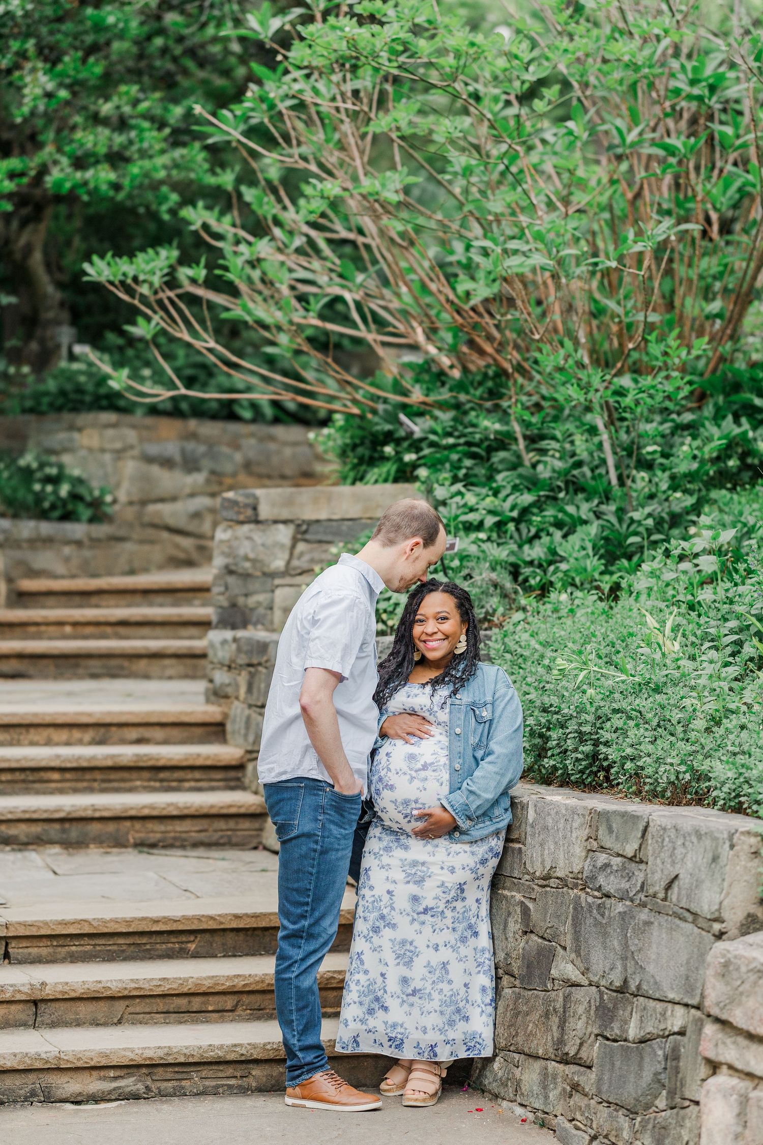 Couple sharing a tender maternity photo moment on stone steps surrounded by lush greenery outdoors.