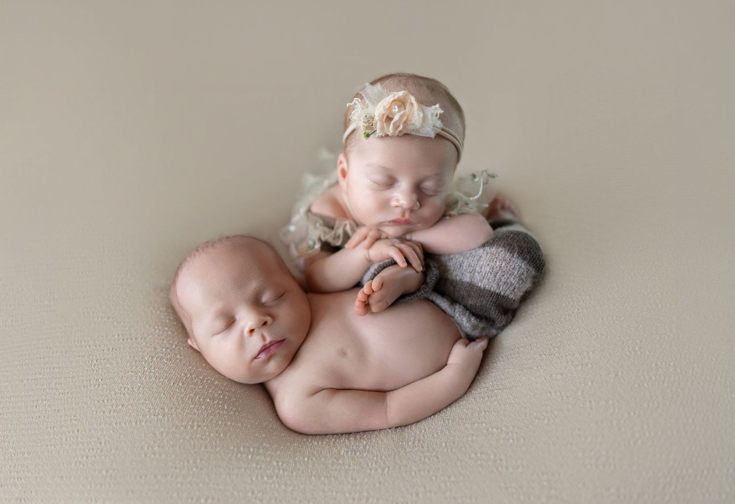 Two sleeping newborn twins posed together on a beige blanket, one wearing a floral headband and lace wrap.
