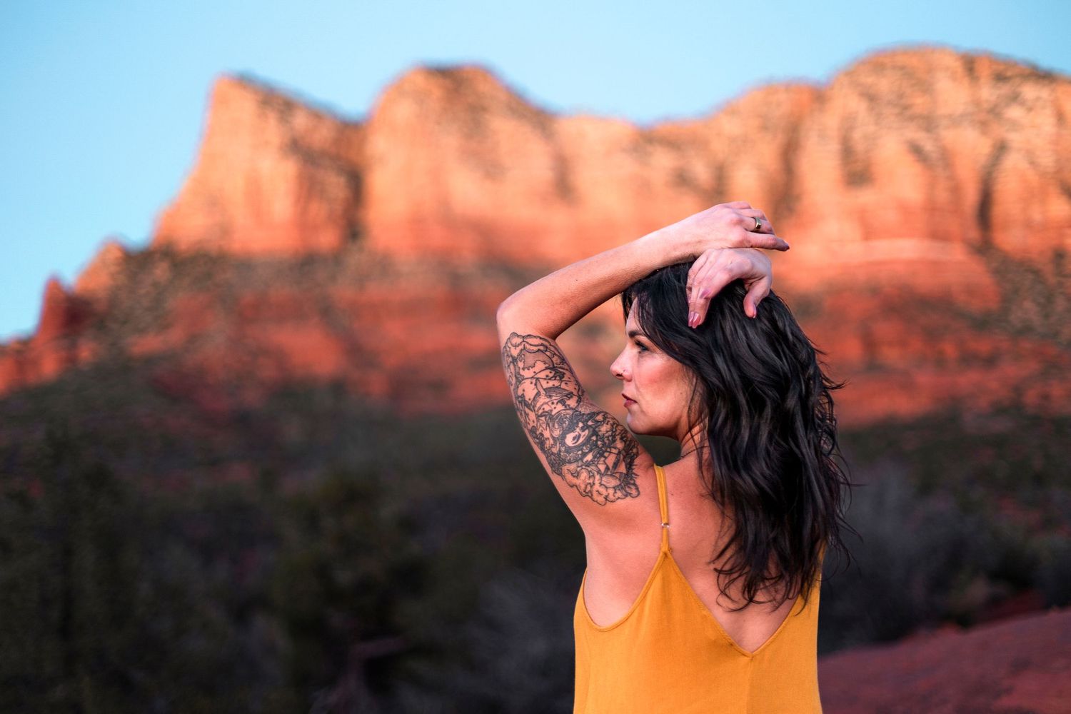 Dark-haired woman with floral arm tattoo in yellow top against Sedona red rock backdrop.