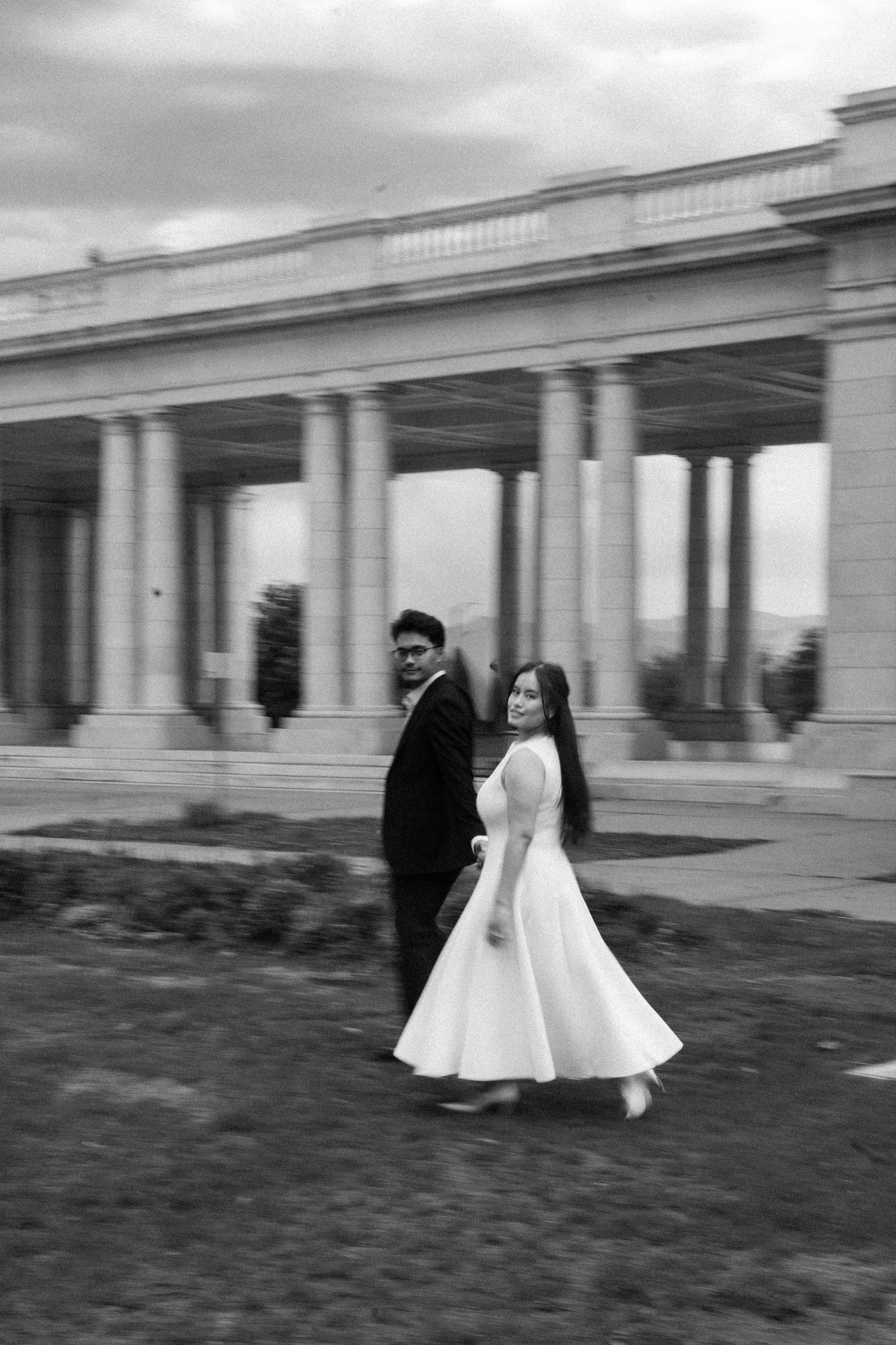 Black and white photo of bride in tea-length dress and groom walking past grand columned architecture.