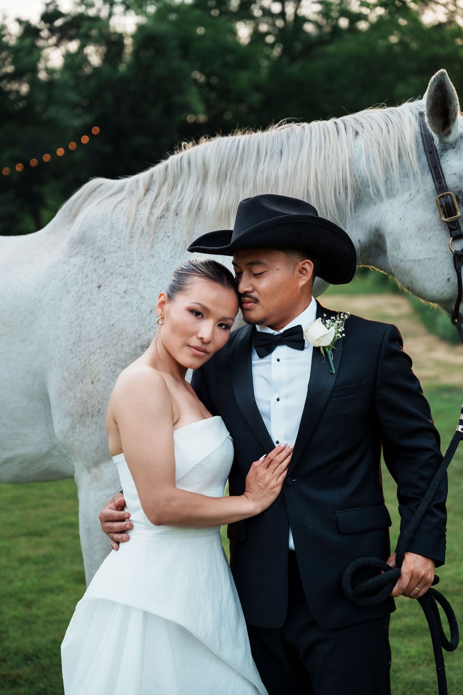 Elegant bride and groom pose beside a white horse, groom wearing a black tuxedo and cowboy hat outdoors.