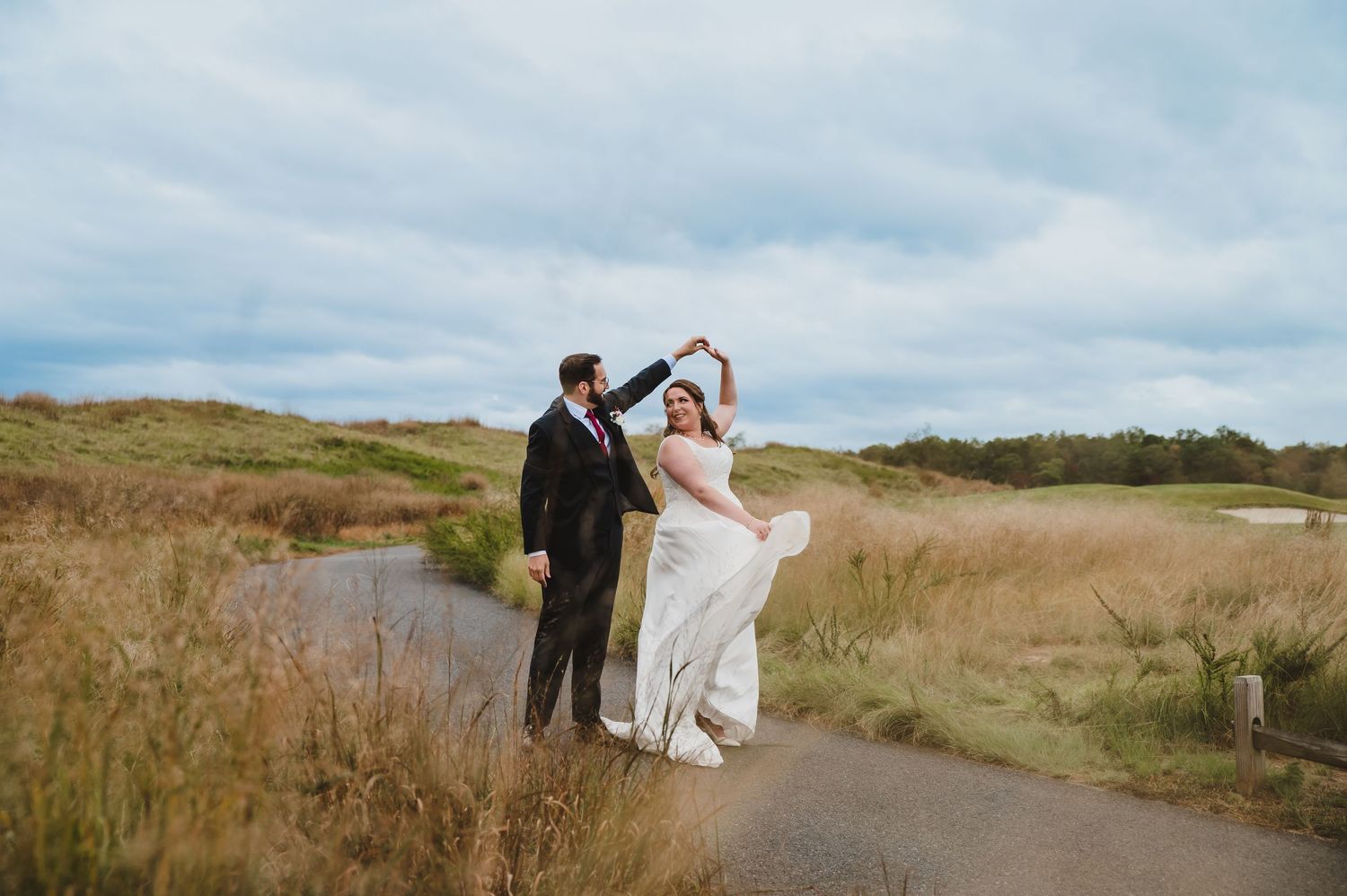 Bride and groom dancing on a path surrounded by golden meadows under a cloudy sky on their wedding day.
