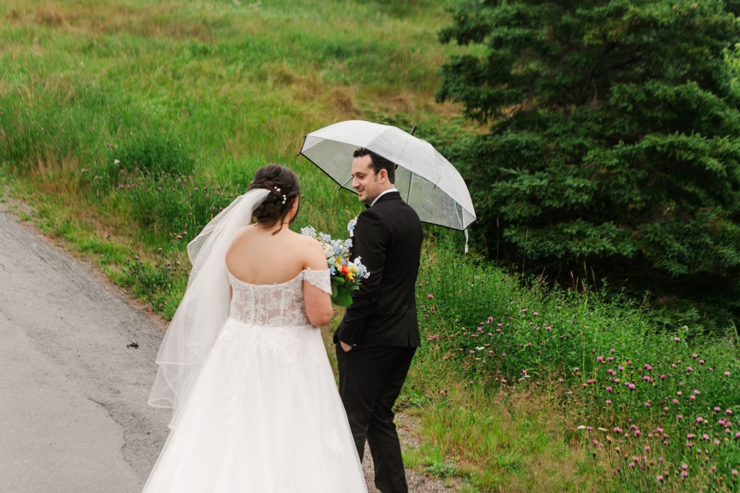 Bride and groom share an umbrella on a rainy wedding day, standing on a path surrounded by lush green grass.