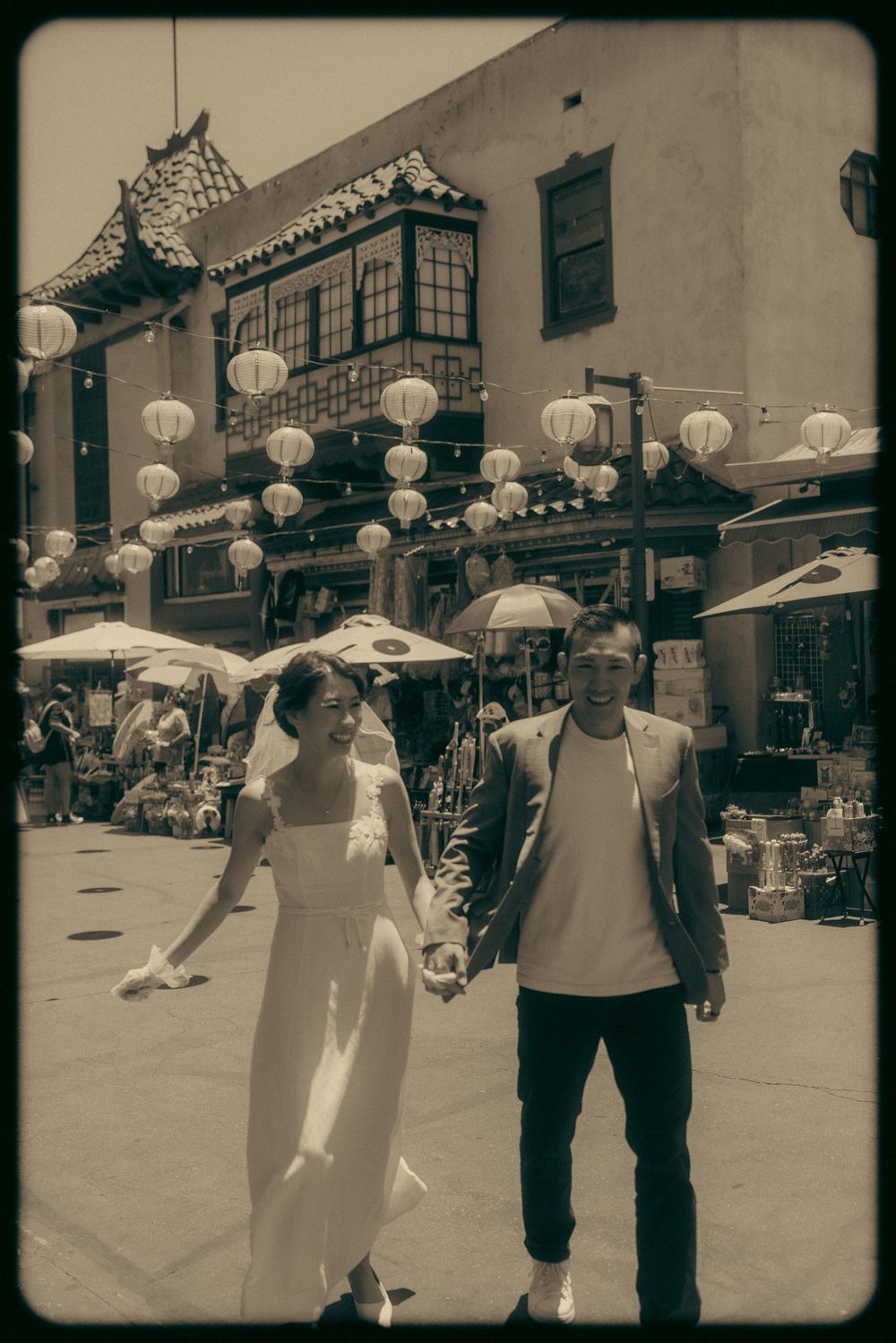 Couple in wedding attire holding hands in Chinatown with white lanterns and traditional architecture in sepia tone.