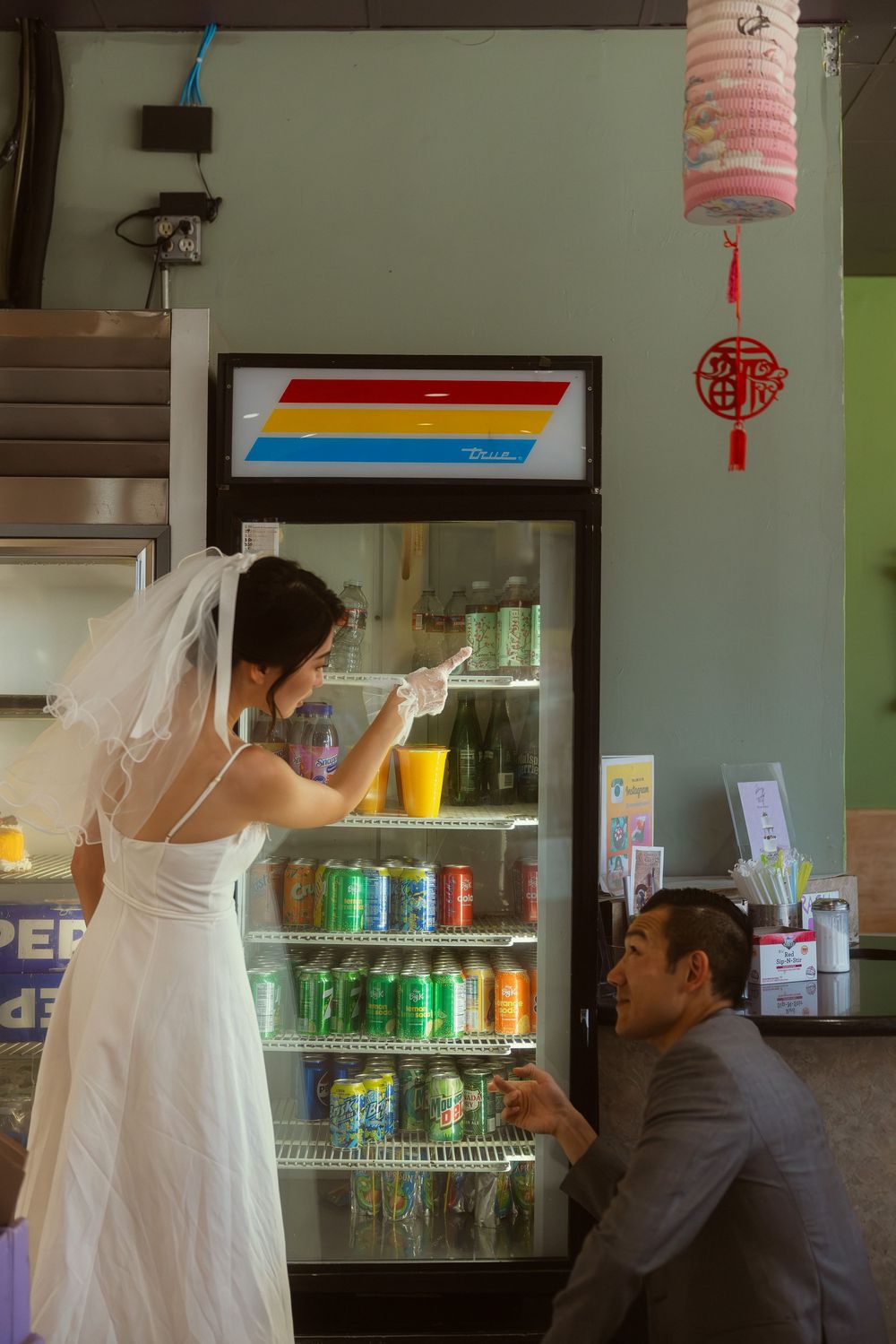 Bride in white wedding dress and veil pointing at drinks in a restaurant refrigerator while groom kneels beside her.