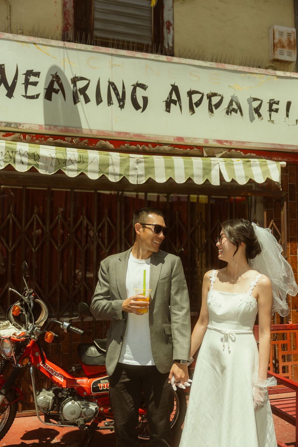 Bride and groom holding hands outside a wearing apparel shop, with a red motorcycle in the background.