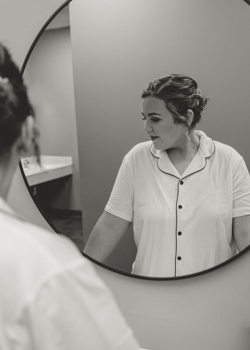 Black and white photo of a woman in white pajamas viewing her reflection in a round mirror while getting ready.